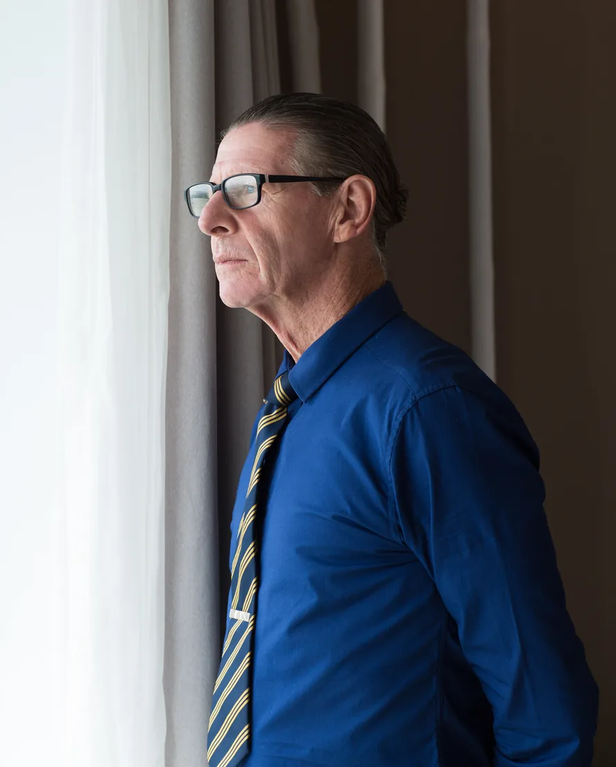 A senior owner in glasses, a blue shirt and a striped tie stands at his office window, looking out, deep in thought