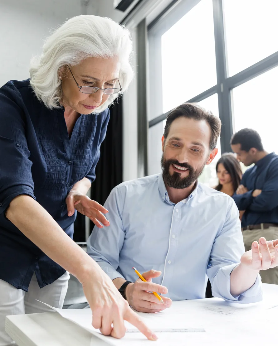 A silver haired owner leans over a desk to point at the plans while a smiling, bearded colleague talks them through, two more colleagues behind