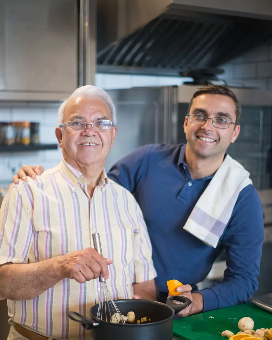 A father and his grown son smile side by side at the stove in their kitchen, the son's hand on his father's shoulder