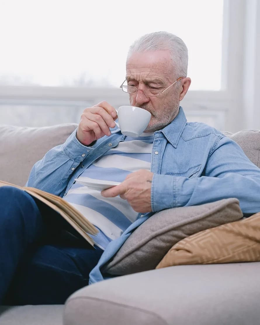 An older man in a denim shirt relaxes on a sofa by a bright window, sipping coffee with a book open on his lap