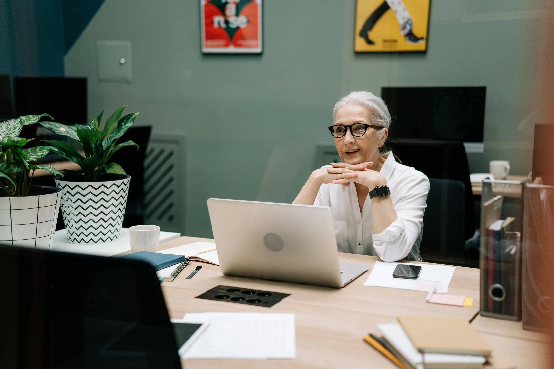 A silver haired businesswoman in glasses smiles at her desk, hands folded behind her laptop, in a warm office