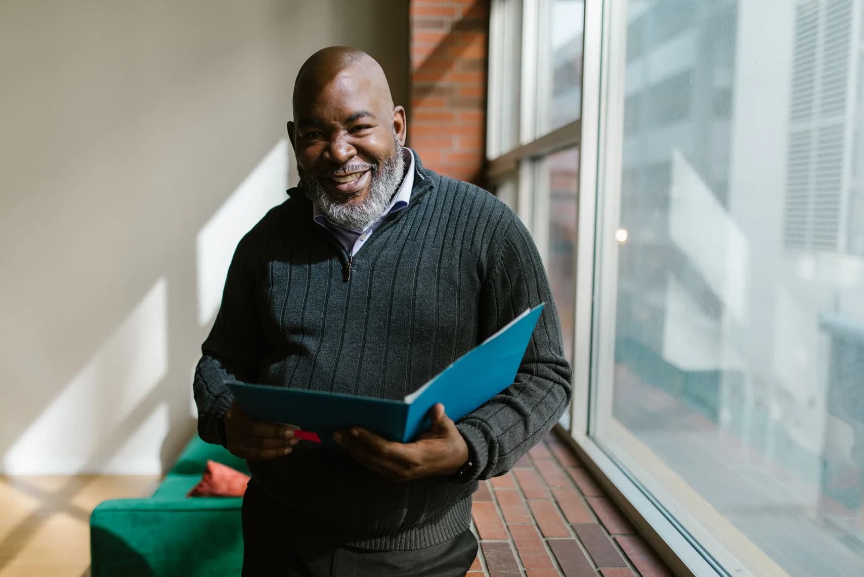 A smiling bearded owner in a dark sweater stands by a sunny window holding an open blue folder